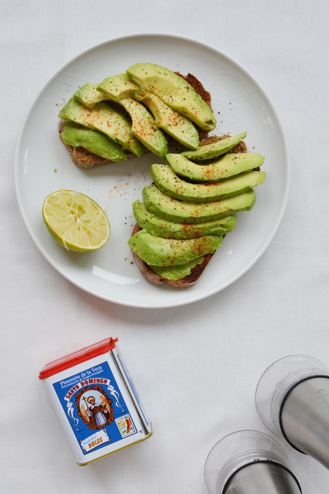 Two pieces of toast topped with avocado and a red seasoning on a white place with a used lime next to a container of Santo Domingo Seasoning and a salt and pepper shaker.
