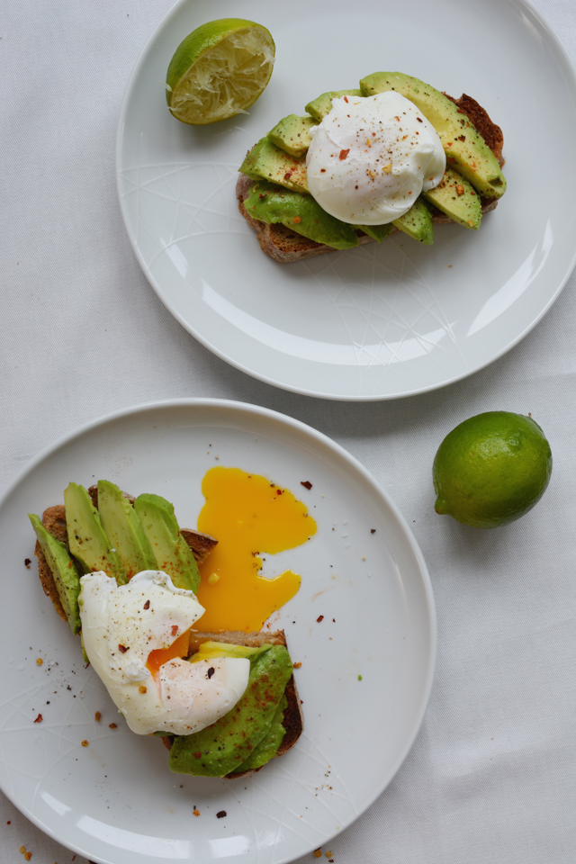 Two white plates with toast topped with avocado and a poached egg next to a lime on a white background.