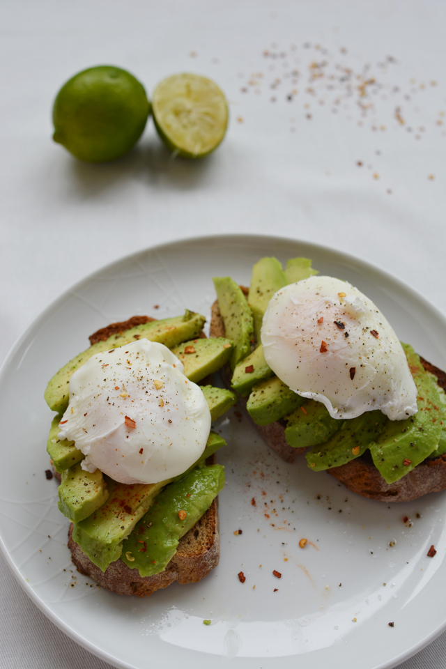 Seasoned avocado topped with a poached eggs on a white plate with a white tabletop with limes in the background.