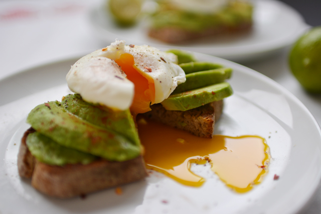 Toast on a white plate topped with sliced avocado and a runny poached egg that is cut in half. Another plate with the same meal is in the background.