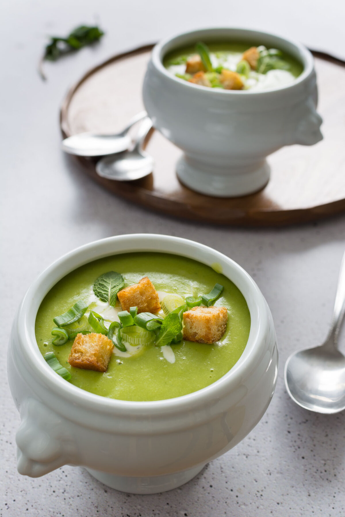Bowl of mint and pea soup with toppings on a white background with another bowl and some spoons in the background on a wooden tray.