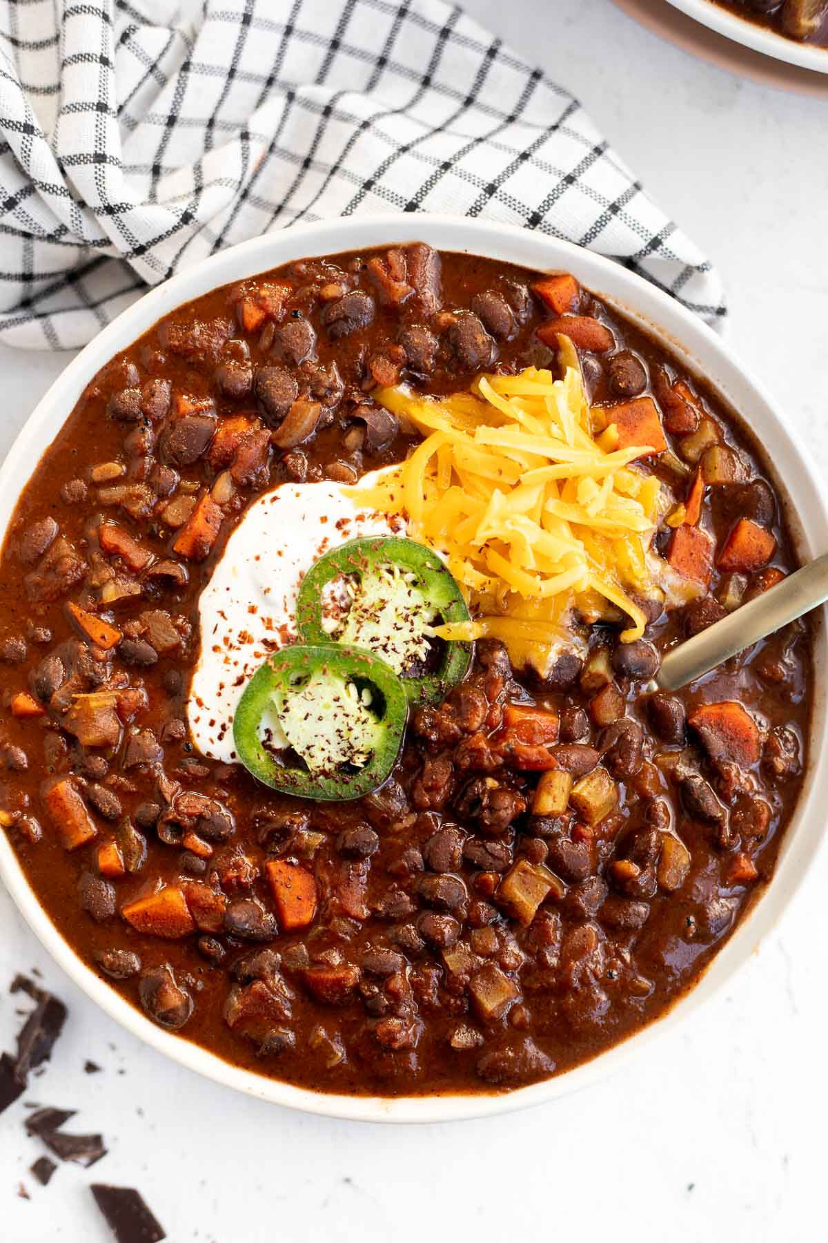 Black Bean Chili with Chocolate in a white bowl garnished with jalapeno slices, sour cream, and shredded cheddar, top view.