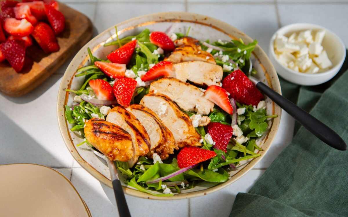 Grilled chicken and strawberry salad with feta cheese, strawberries, and a green cloth napkin in the background on a white tile table.