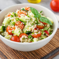 Couscous salad with fresh vegetables and herbs in a bowl on a gray concrete background.