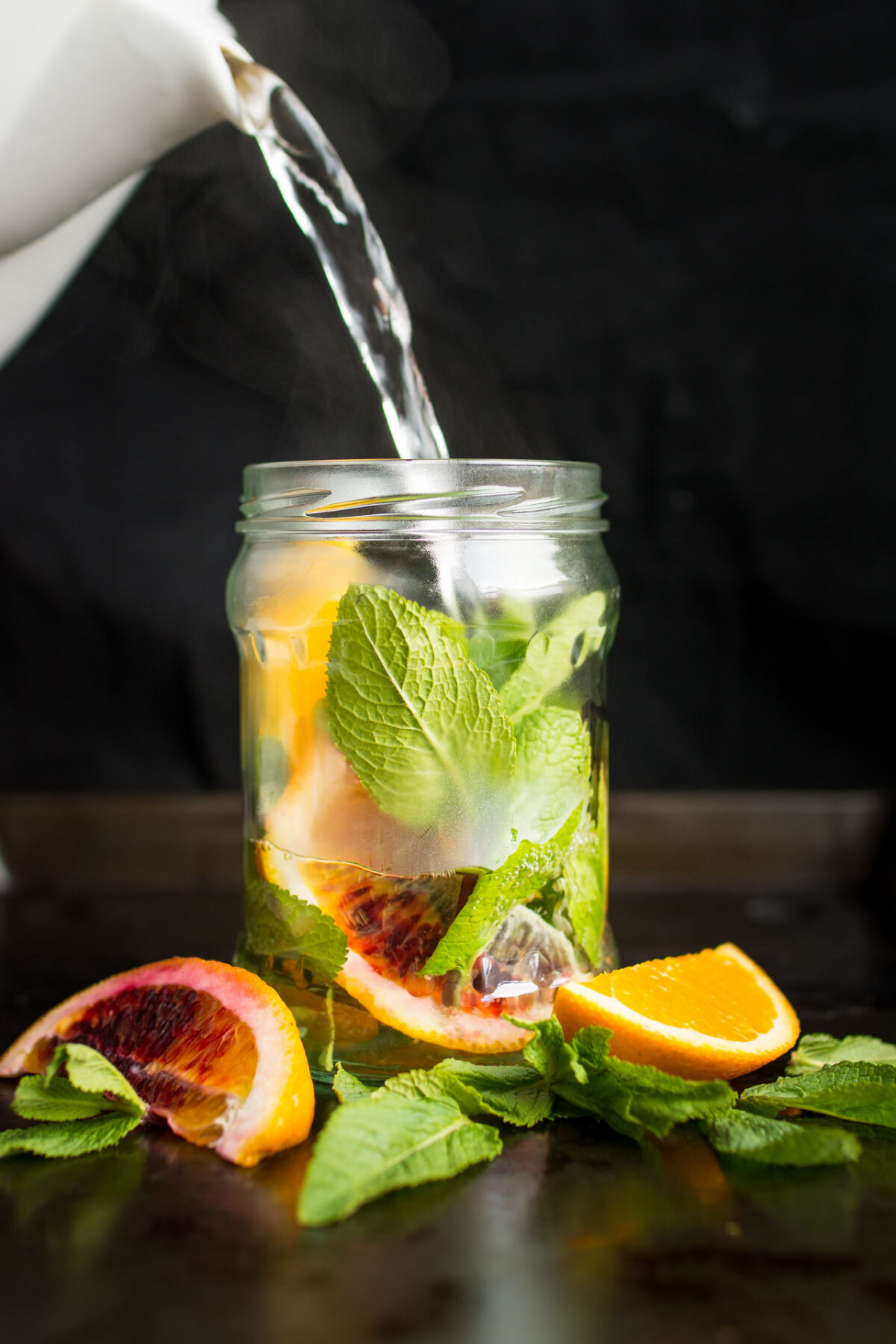 A pitcher of hot water being poured into a glass jar containing sliced oranges and fresh mint sprigs.