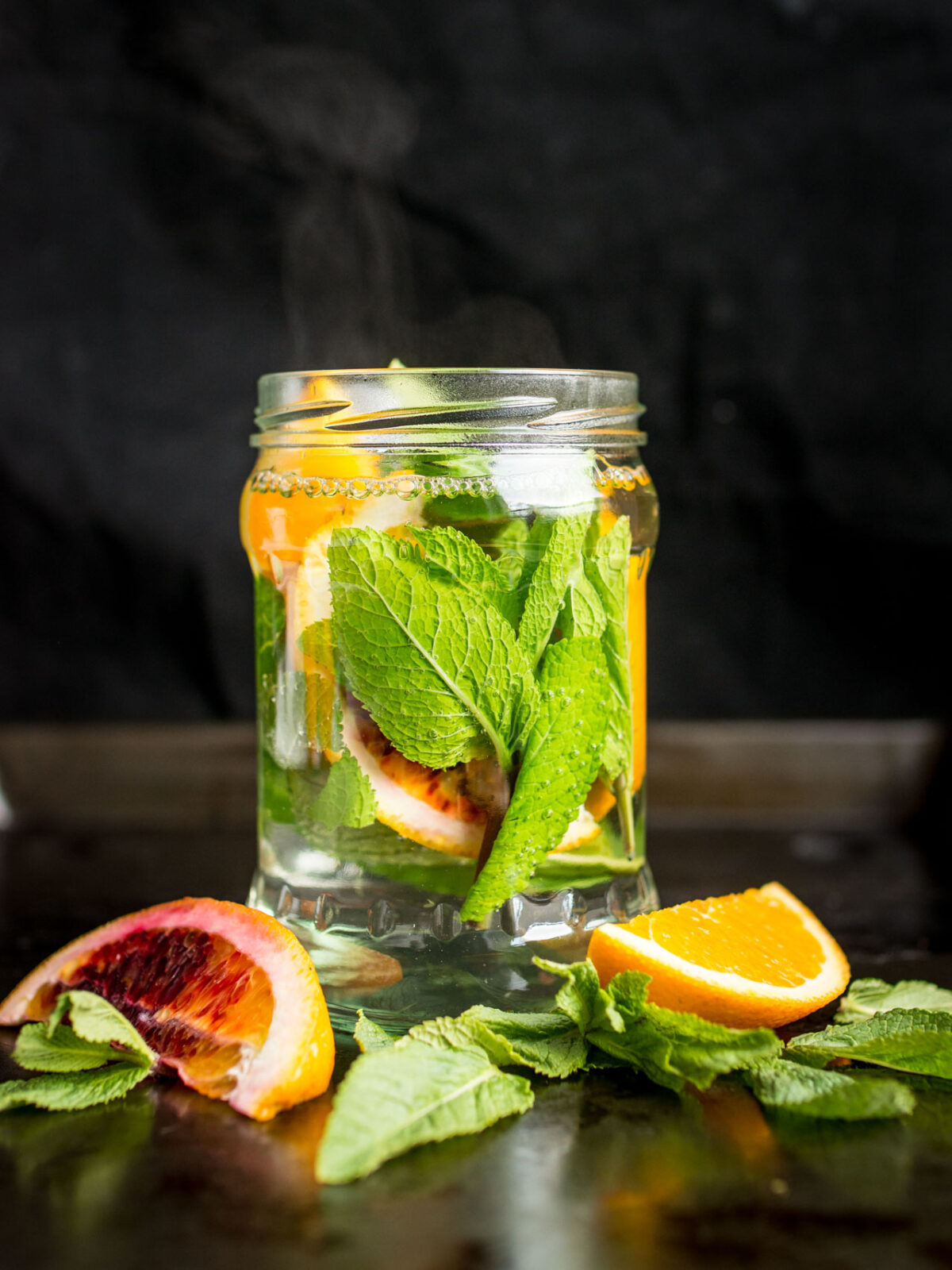 A glass jar containing sliced oranges and fresh mint sprigs floating in hot water to create a refreshing tea.
