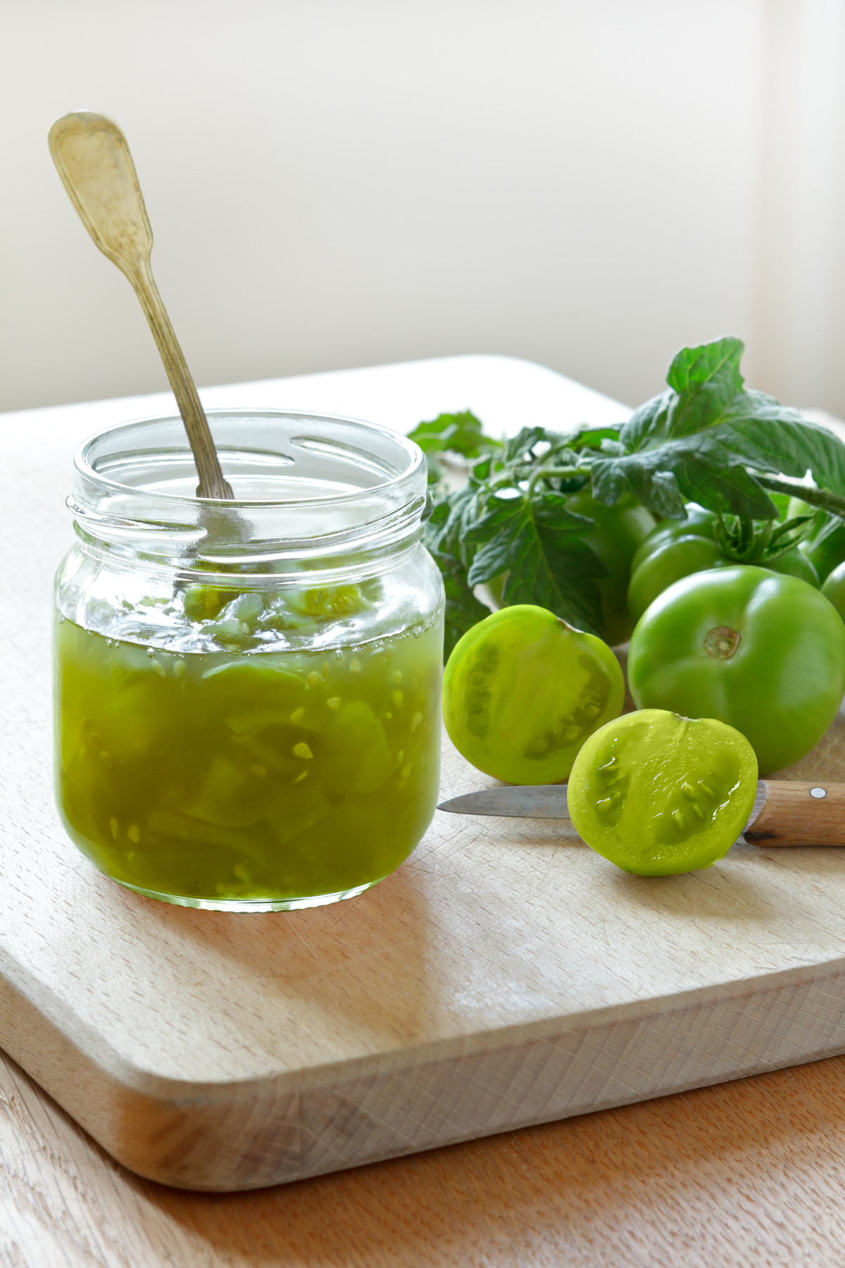 Green tomato chutney in a glass jar on a cutting board with a spoon and green tomatoes in the background.