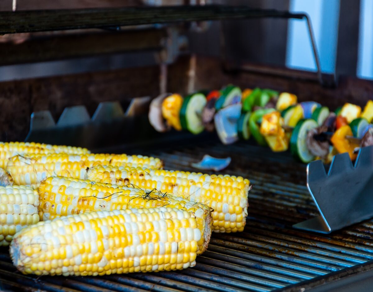 Several ears of corn on a BBQ grill with kabobs in the background.
