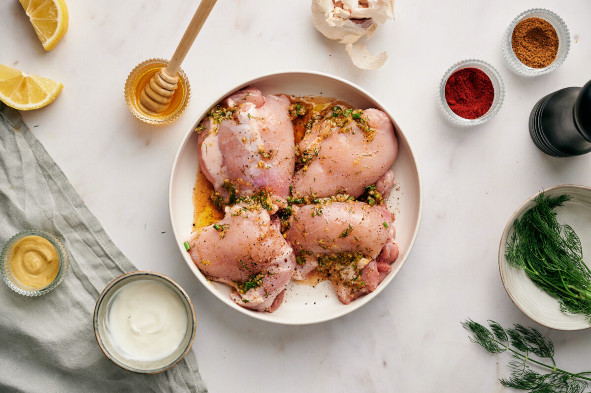 Chicken thighs on a white plate being marinated with lemon and dill seasonings.