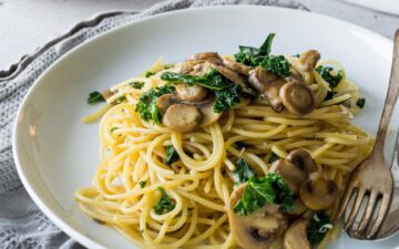 Sauted mushrooms and kale over the top of cooked pasta on a white plate and cloth napkin with a fork and spoon for serving.