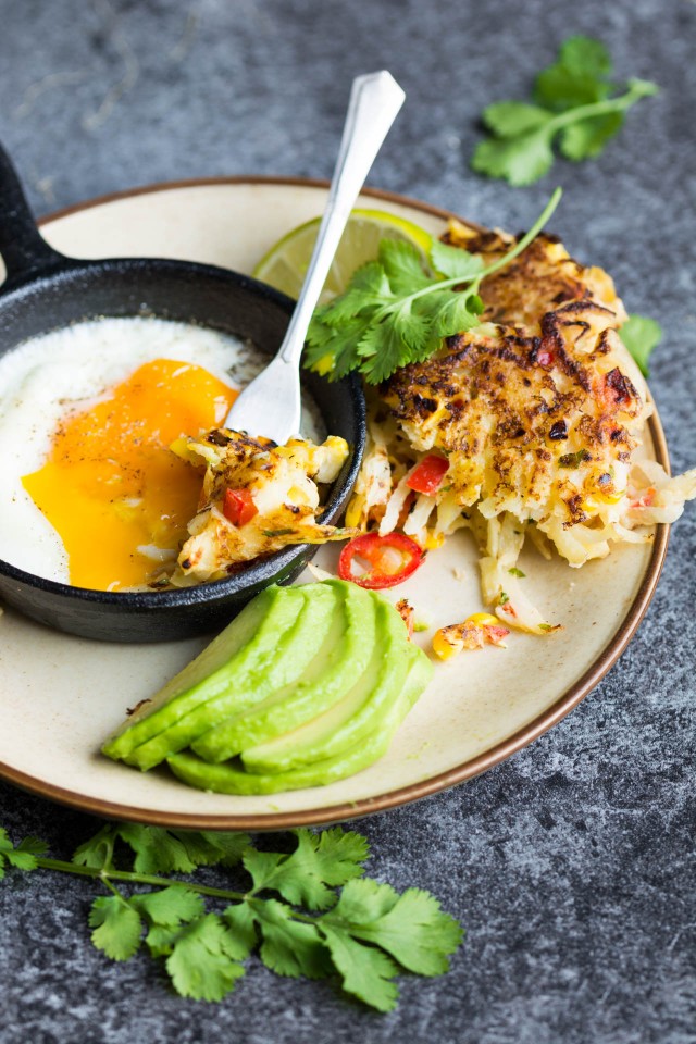 An appetizing photo of a fork tossing a flavorful spicy sweetcorn and potato fritter onto a plate, accompanied by sliced avocado and a perfectly cooked sunny-side-up egg.