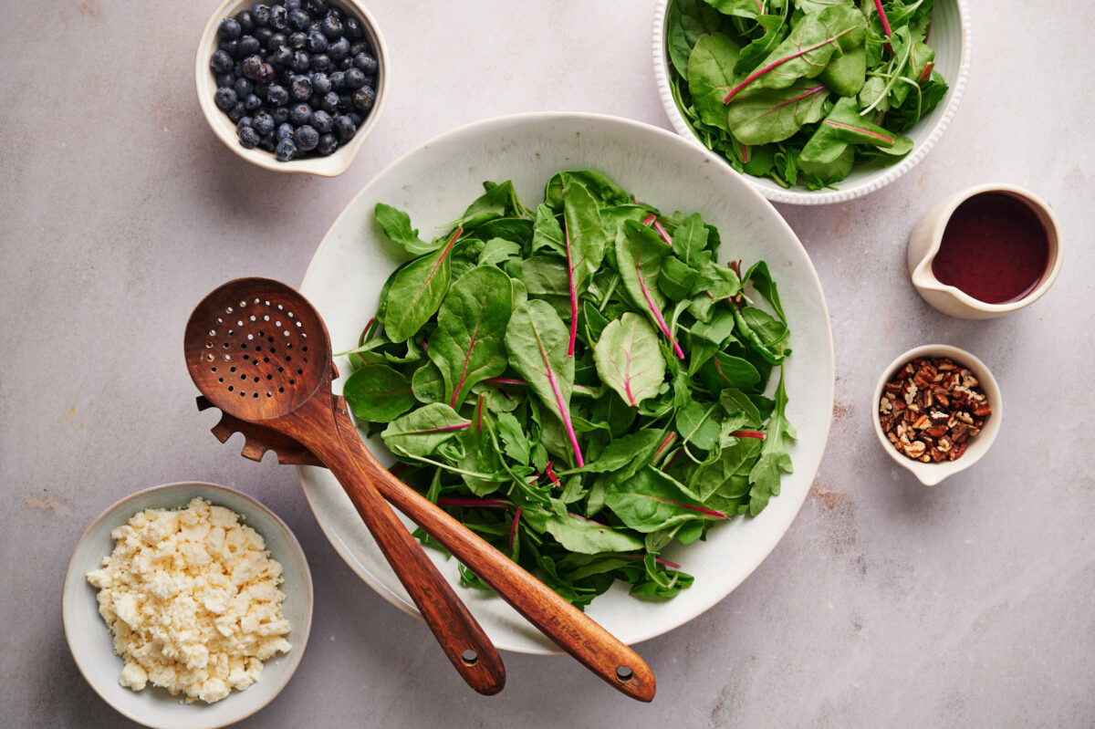 Spring mix (baby spinach and arugula) in a white bowl with ingredients for a Summer Blueberry Salad.