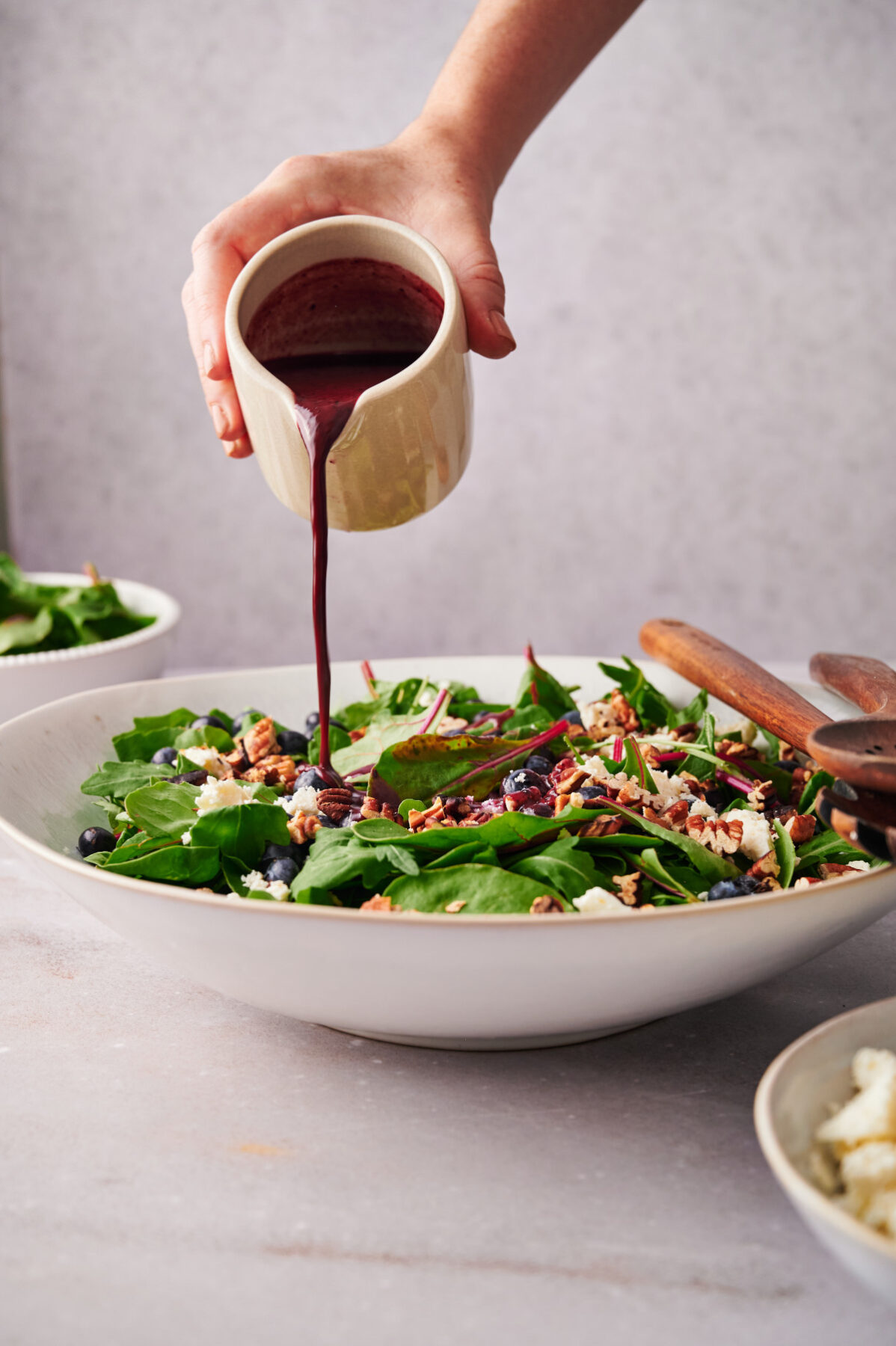 Vertical view of woman's hand pouring blueberry vinaigrette on top of a summer blueberry salad in a white bowl.