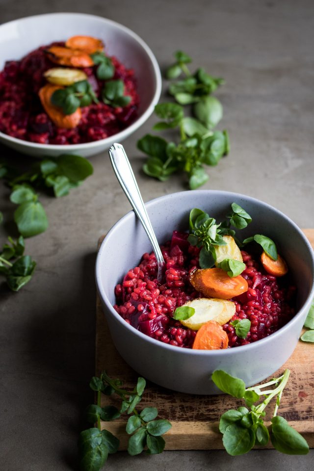 Side view of a red beet and barley risotto served with roasted carrots in white bowls.