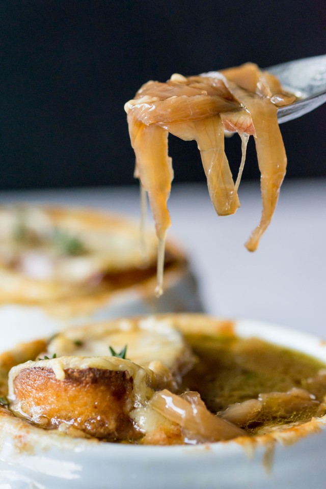 Spoonful of French Onion Soup with onions on it being held above a bowl of French Onion Soup in a white bowl topped with bread and cheese.