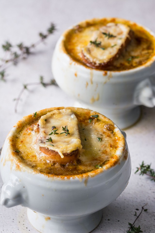 Two bowls of Vegetarian French Onion soup topped with bread and cheese on a white table sprinkled with herbs.