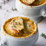 Vegetarian French Onion soup in a white bowl on a white table with herbs on the table and another bowl of soup in the background.